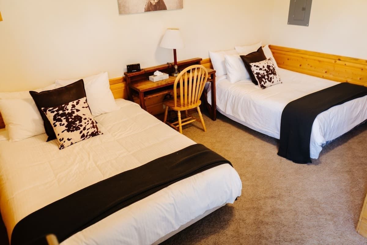 Two beds in a room, each with a dark throw and cow-print pillow. Desk and chair between them.