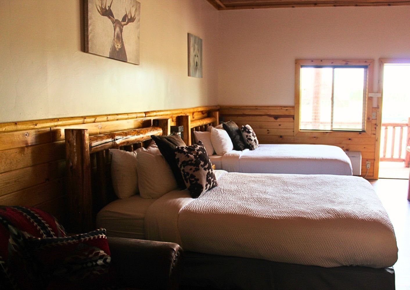 Two beds in a rustic wood-paneled hotel room, with a small seating area and window.
