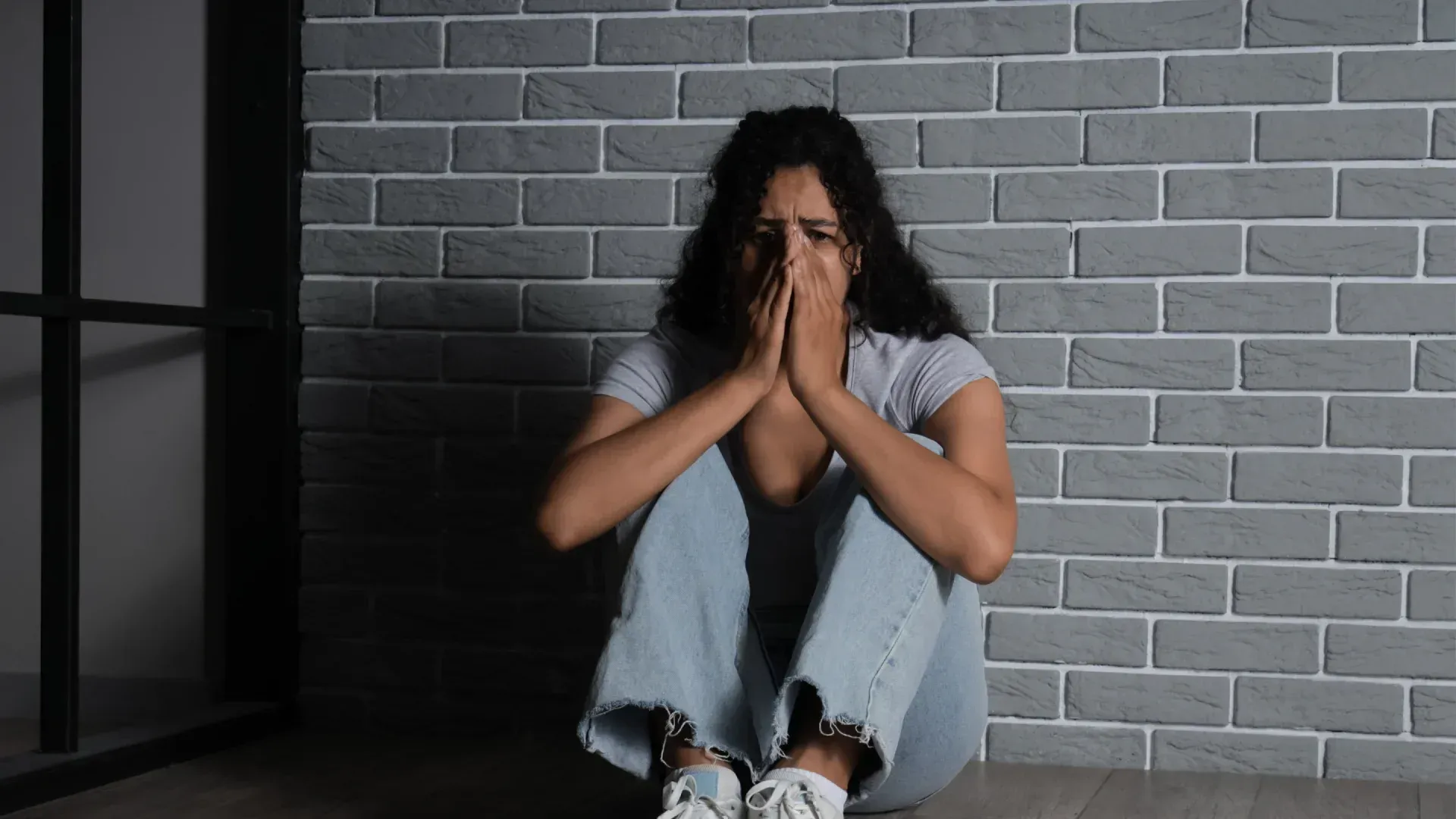 Woman sits on floor, crying with hands covering face, against gray brick wall.
