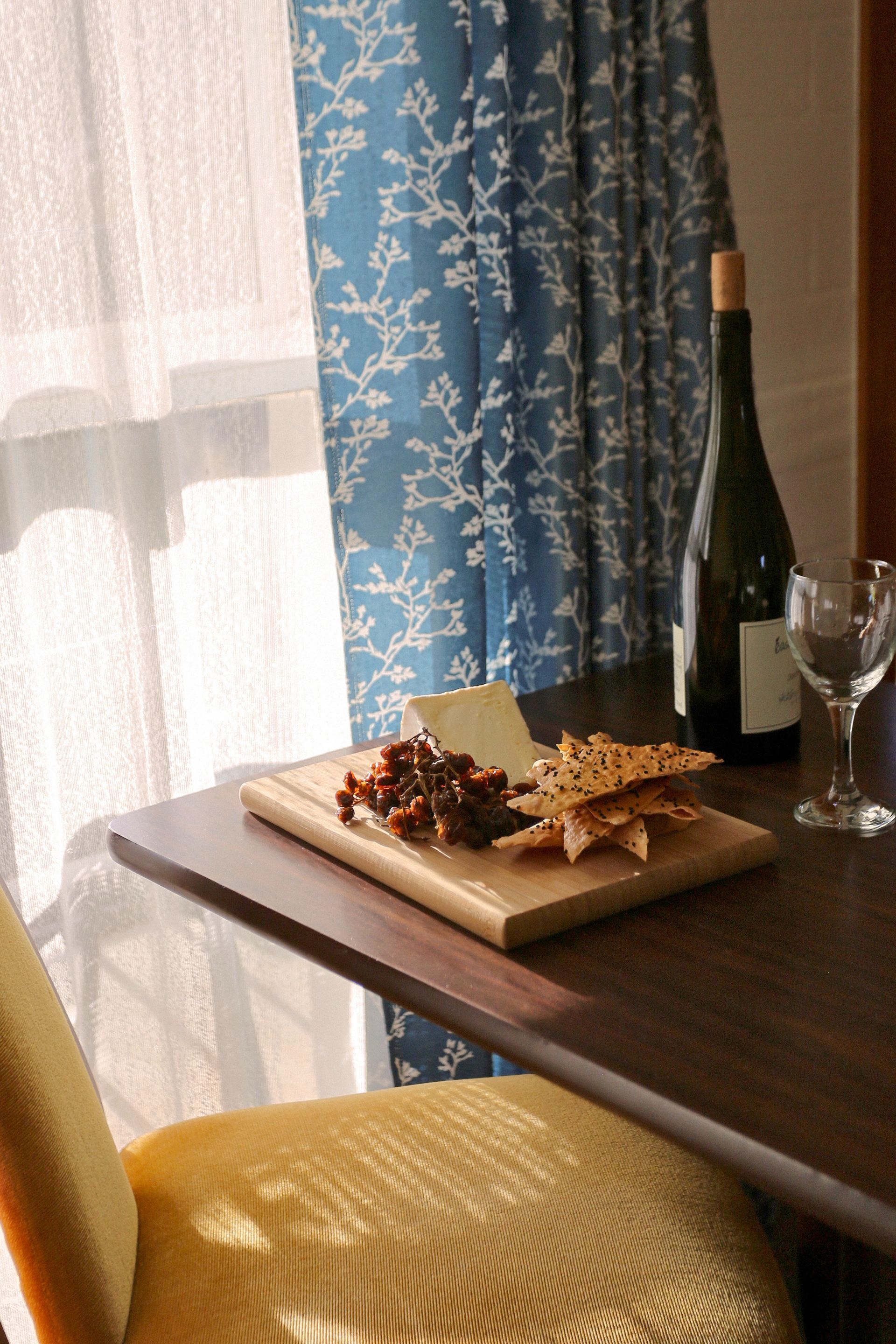 Cheese and crackers on a wooden board next to wine and a glass on a table in front of a blue curtain.