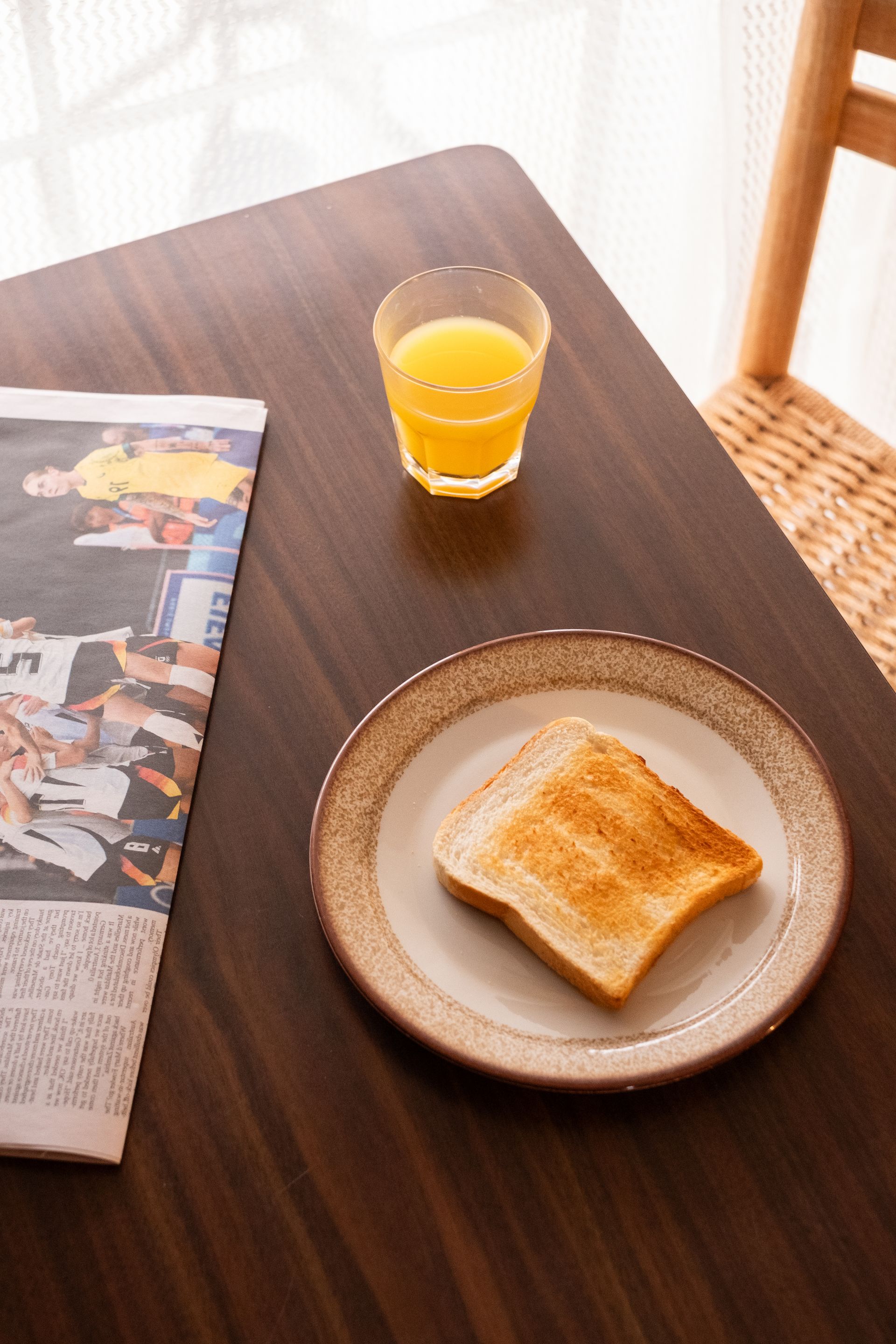 Toast on a plate, orange juice in a glass, and a newspaper on a dark wood table near a window.