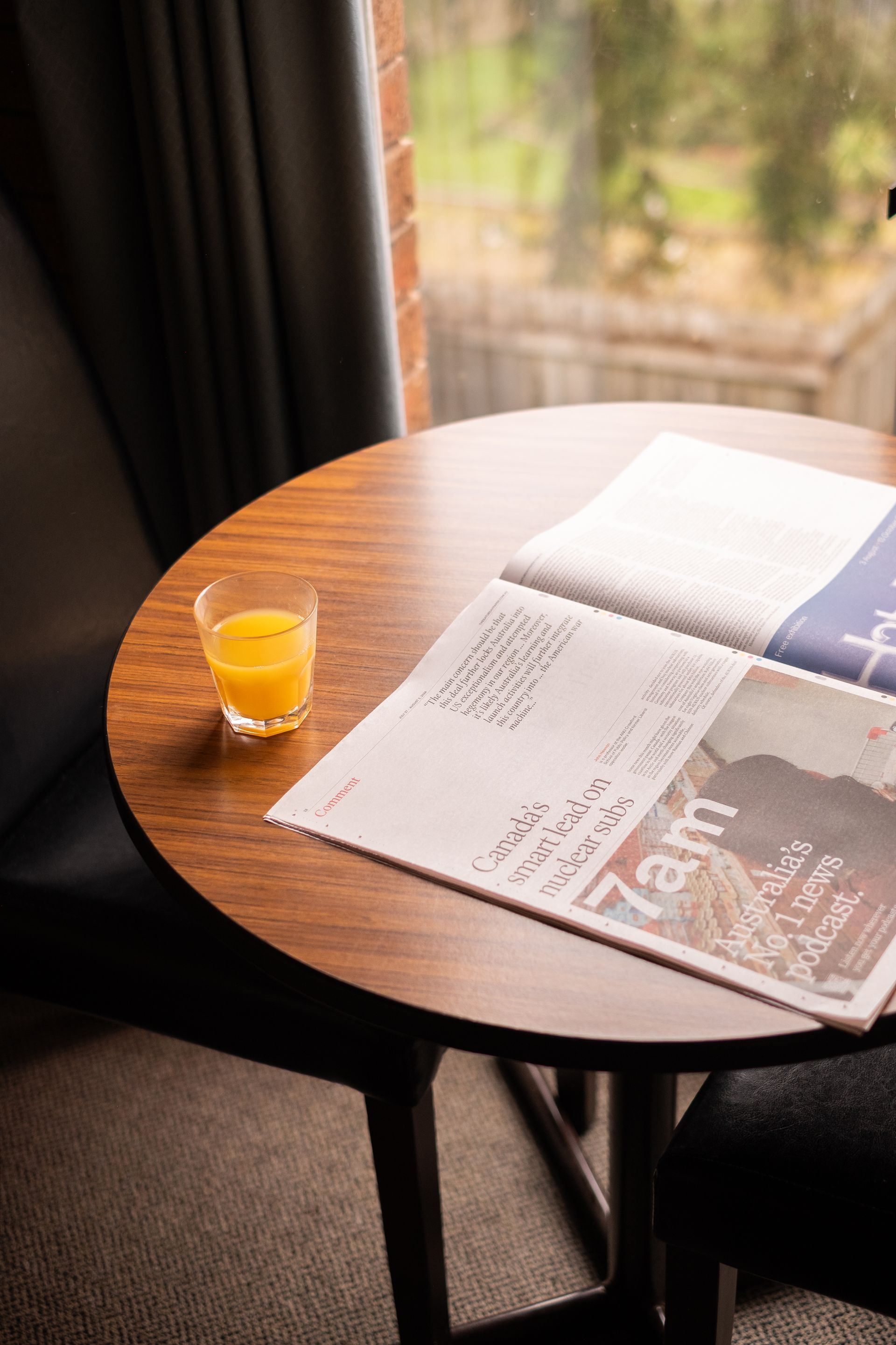 Round wooden table with newspaper and orange juice in front of a window, next to a chair.