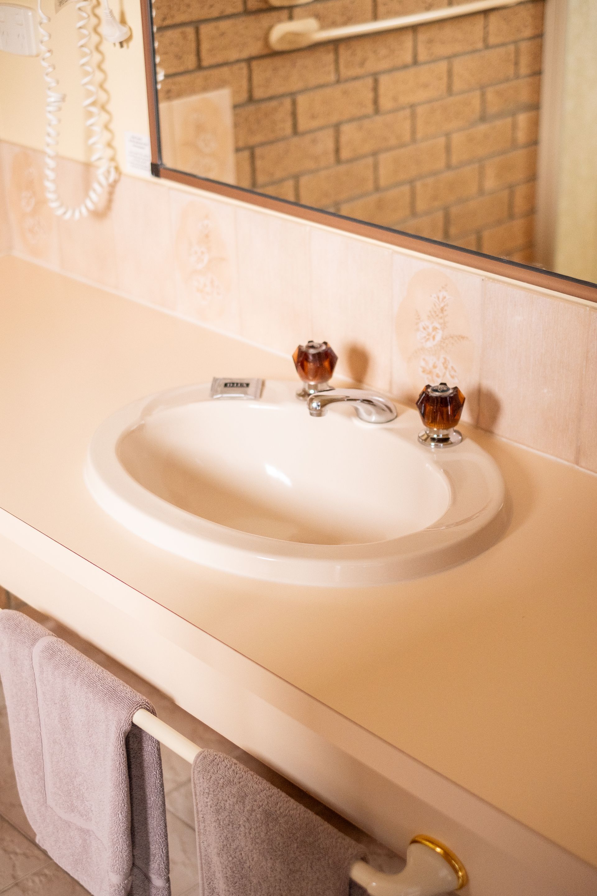 Bathroom sink with cream countertop, brick wall mirror, and towels.