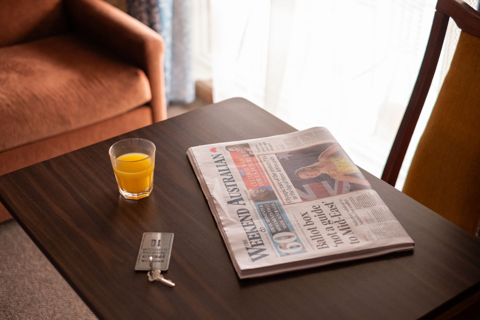 Table with newspaper, glass of juice, and medication next to an armchair and window.