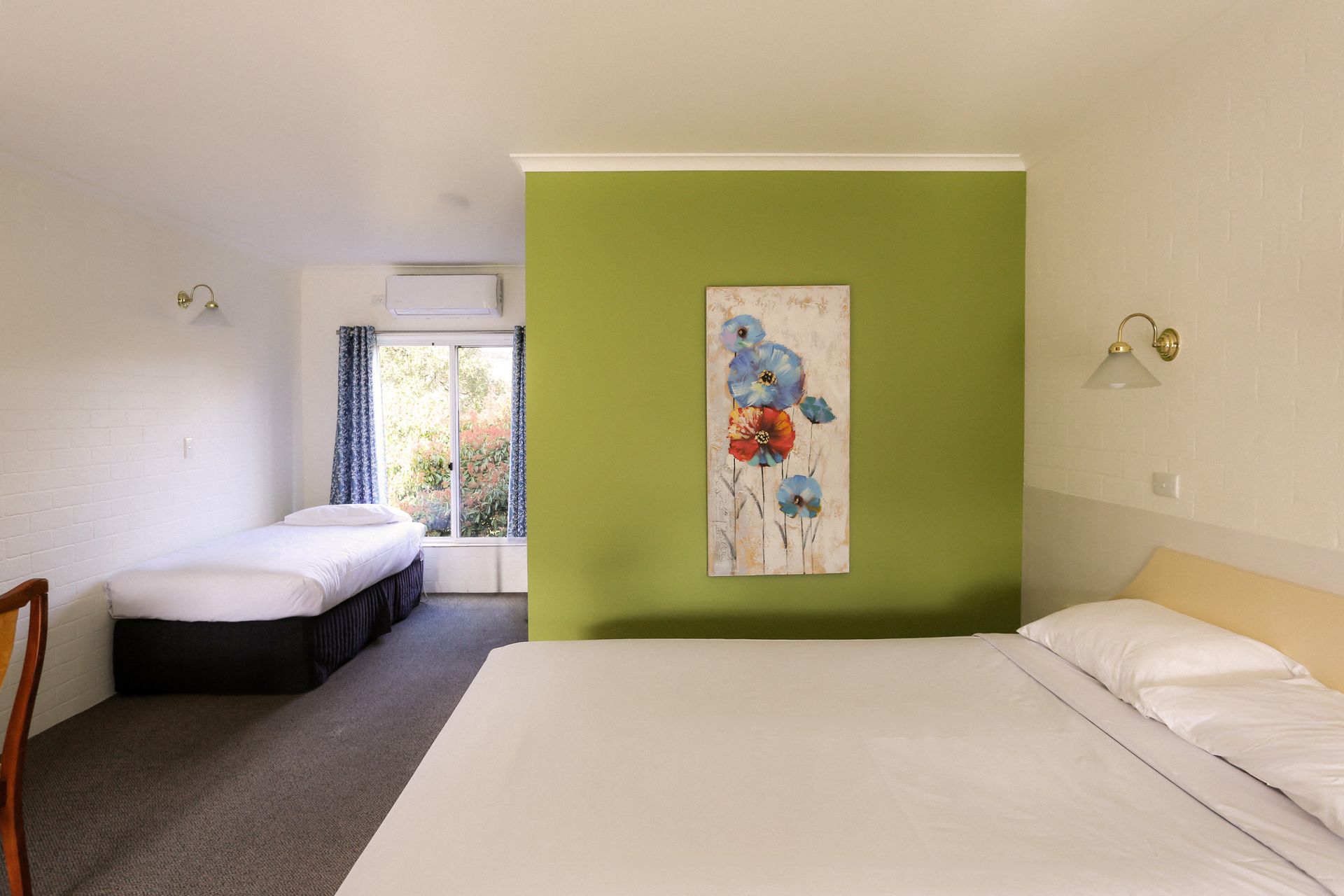 Hotel room with a double bed, single bed, olive green accent wall, and window overlooking trees.