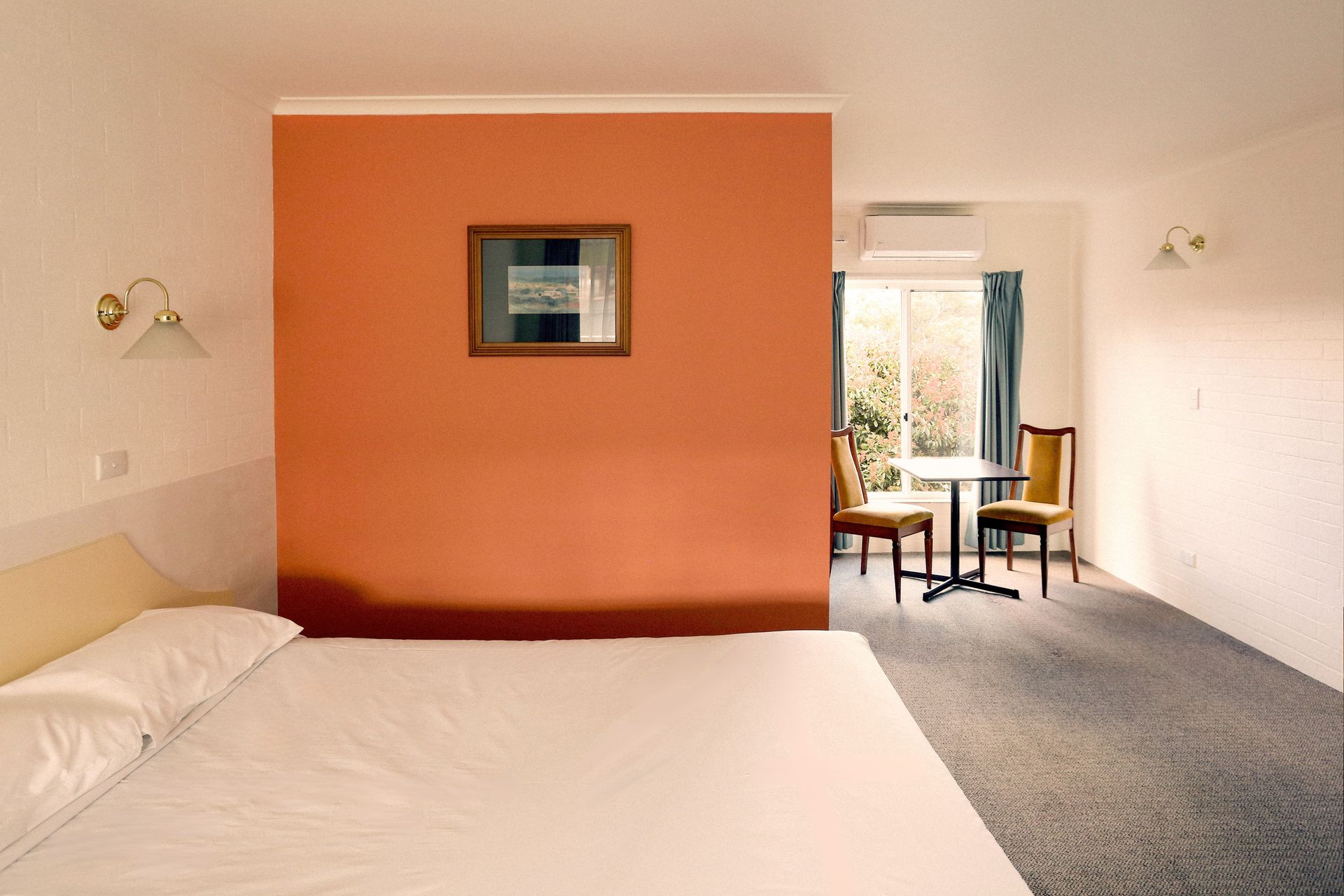 Hotel room with bed, orange wall, small table, and chairs near a window.