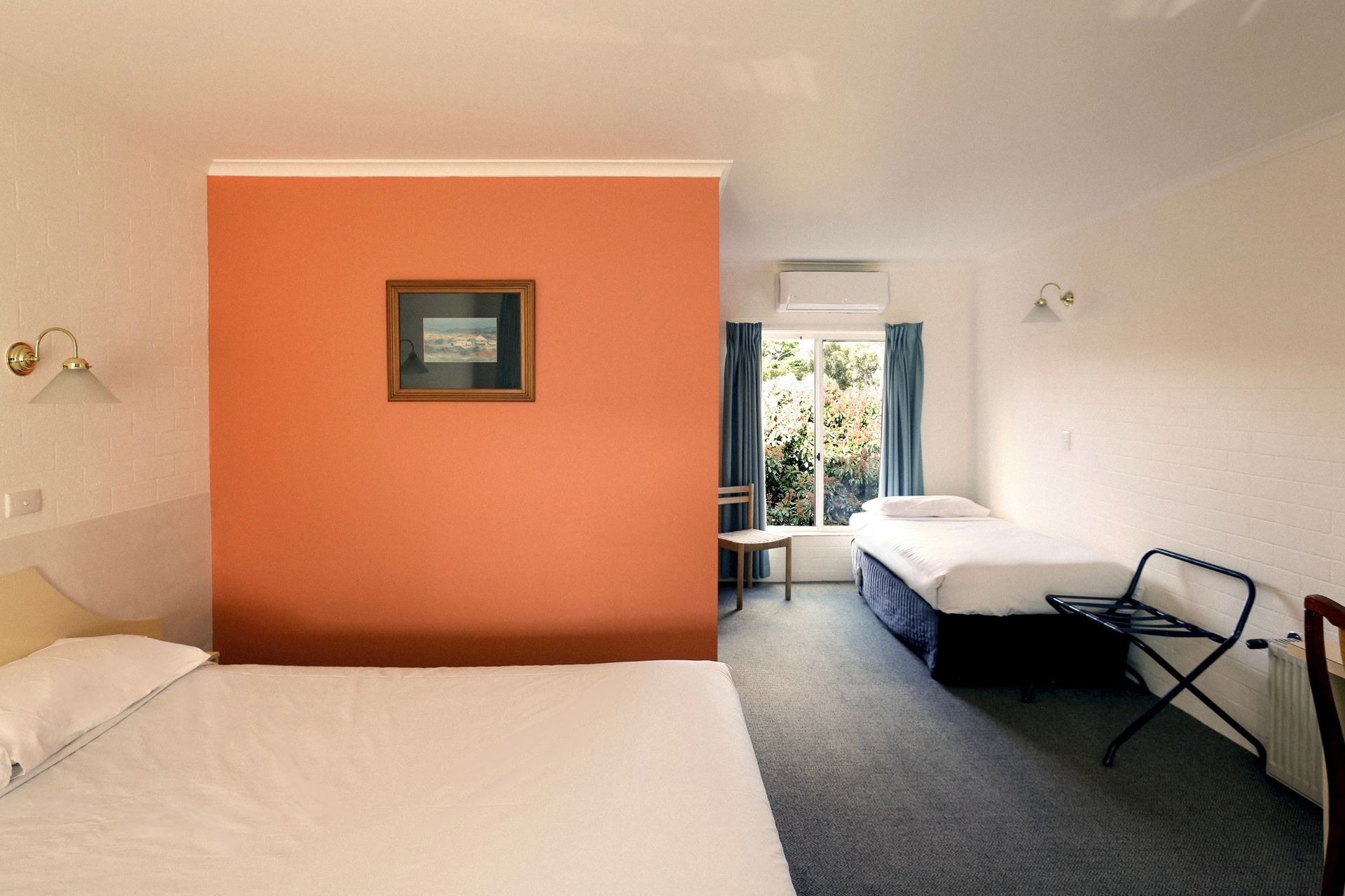 Hotel room with orange accent wall, bed, single bed, window with curtains, and luggage rack.
