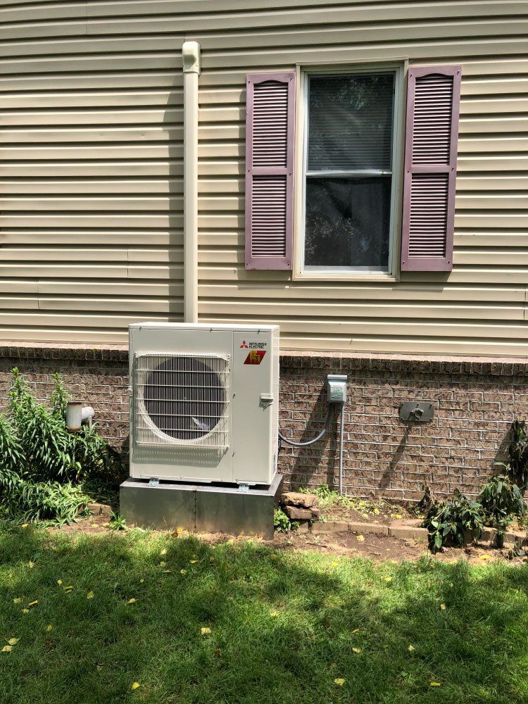 A white Mitsubishi heat pump unit outside a home, next to a window with purple shutters.