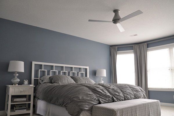 Bedroom with a blue wall, white bed, and ceiling fan; large windows with drapes, and a bench at the foot of the bed.