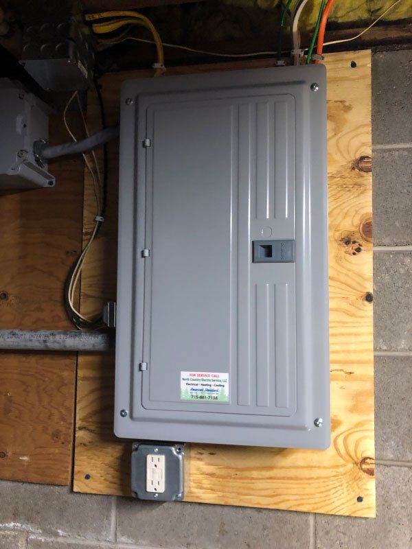 A gray electrical panel mounted on plywood, with an electrical outlet below, against a concrete wall.