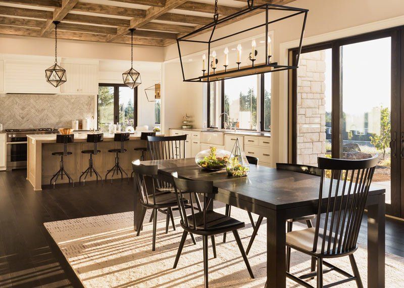 Open-concept dining room with dark wood table, black chairs, and kitchen island. Large windows and modern light fixtures.