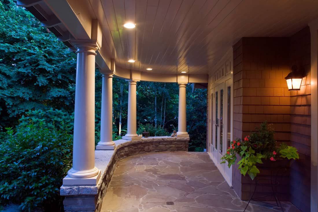 Covered porch with columns, lit by recessed lights and a wall sconce; opens to a wooded area.