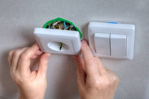Hands installing a white electrical outlet cover, next to a double light switch on a gray wall.
