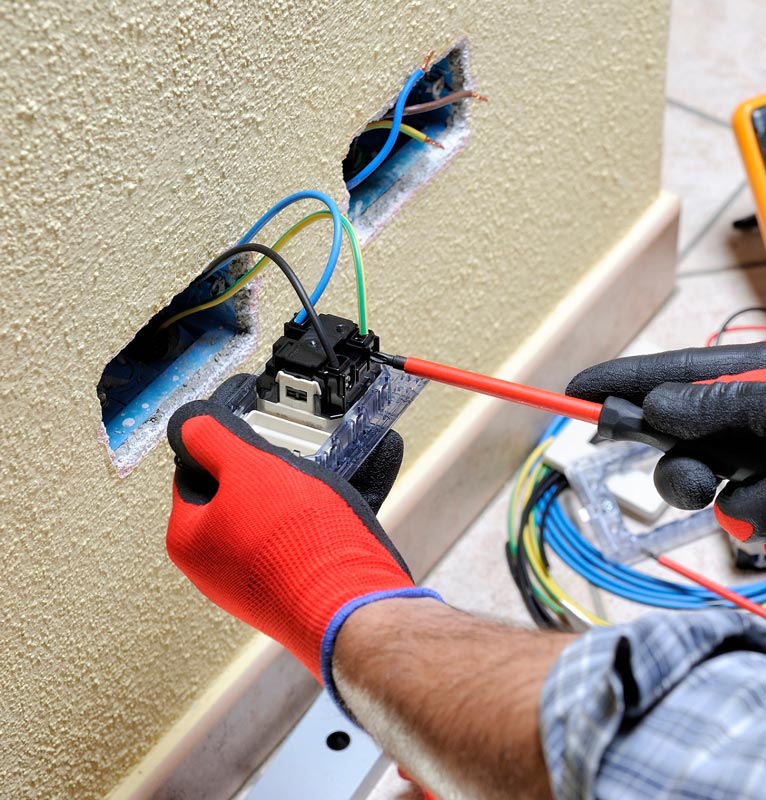 Electrician installing an outlet, wearing gloves, working on a wall with exposed wires, using a screwdriver.