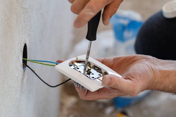 Person installing an electrical outlet, holding it with one hand and using a screwdriver with the other. Wires are visible.