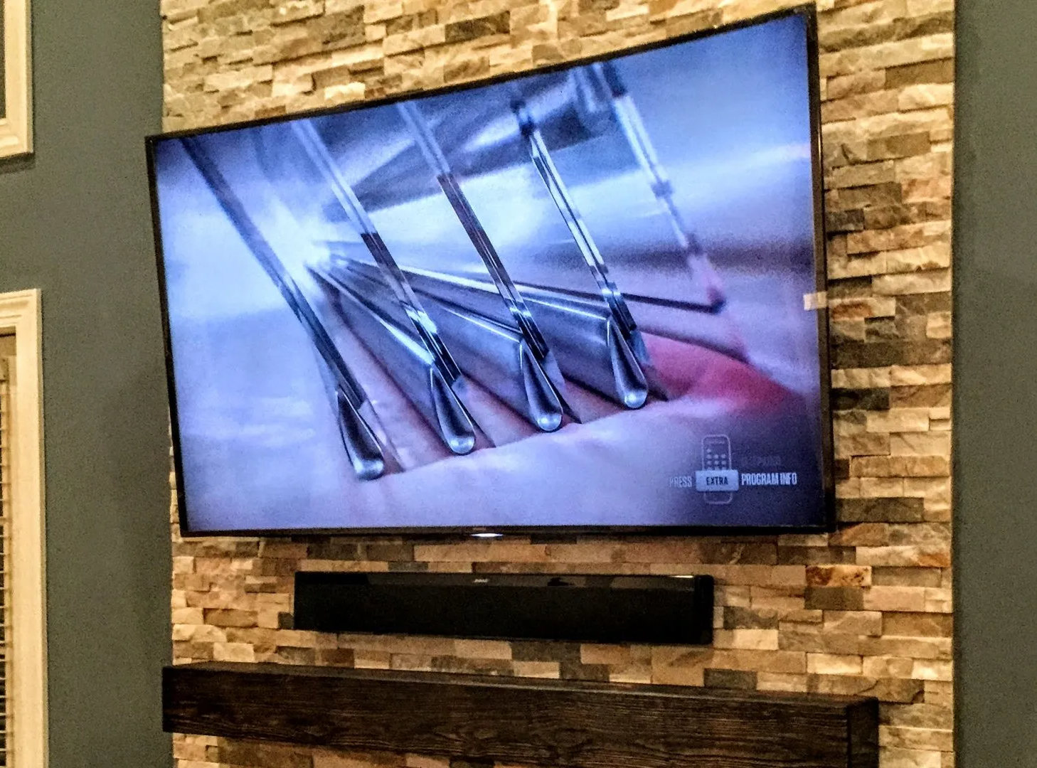 Curved TV mounted on a stone wall, above a black soundbar and wooden shelf.