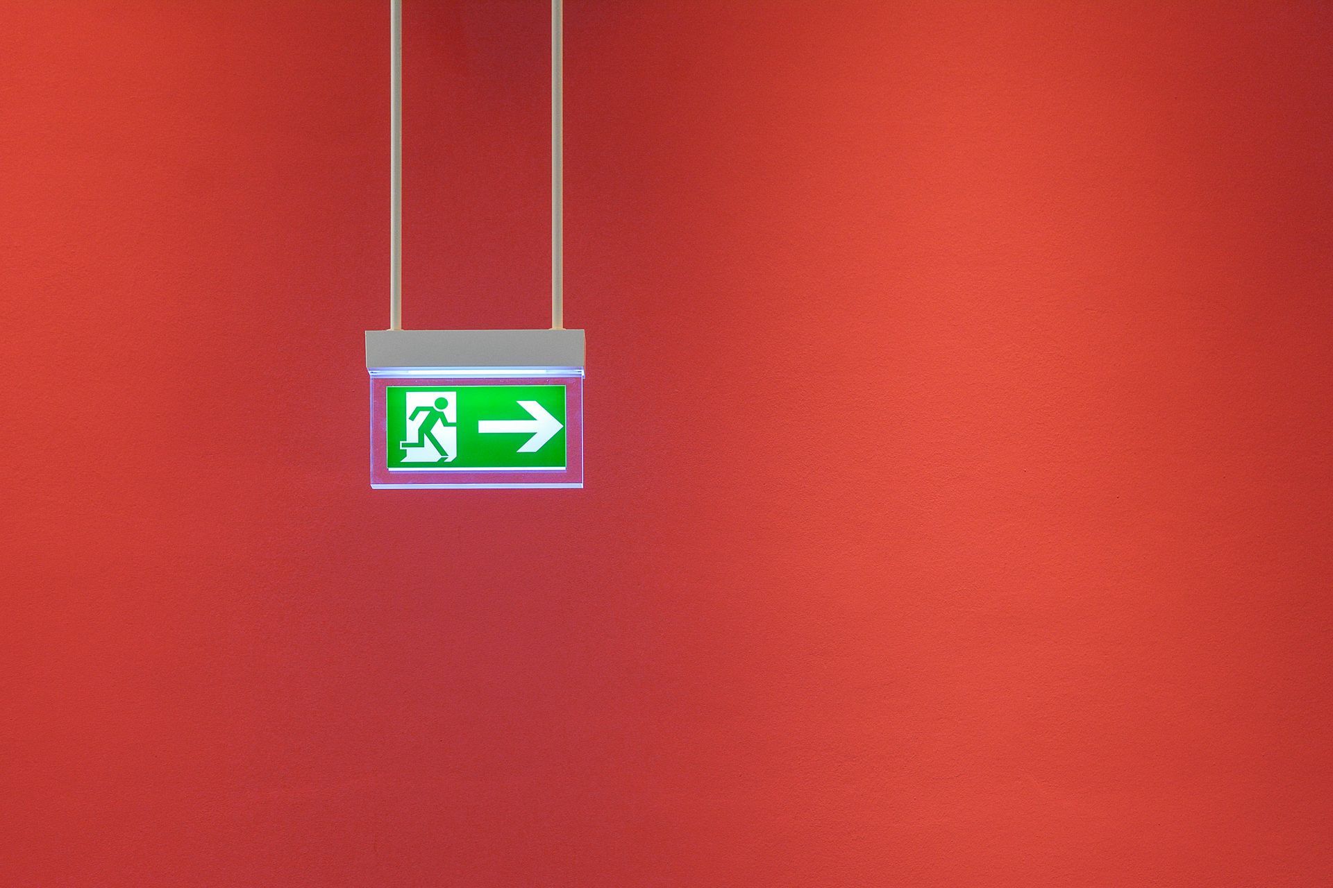 Green exit sign pointing right, hanging from white cords against a red wall.