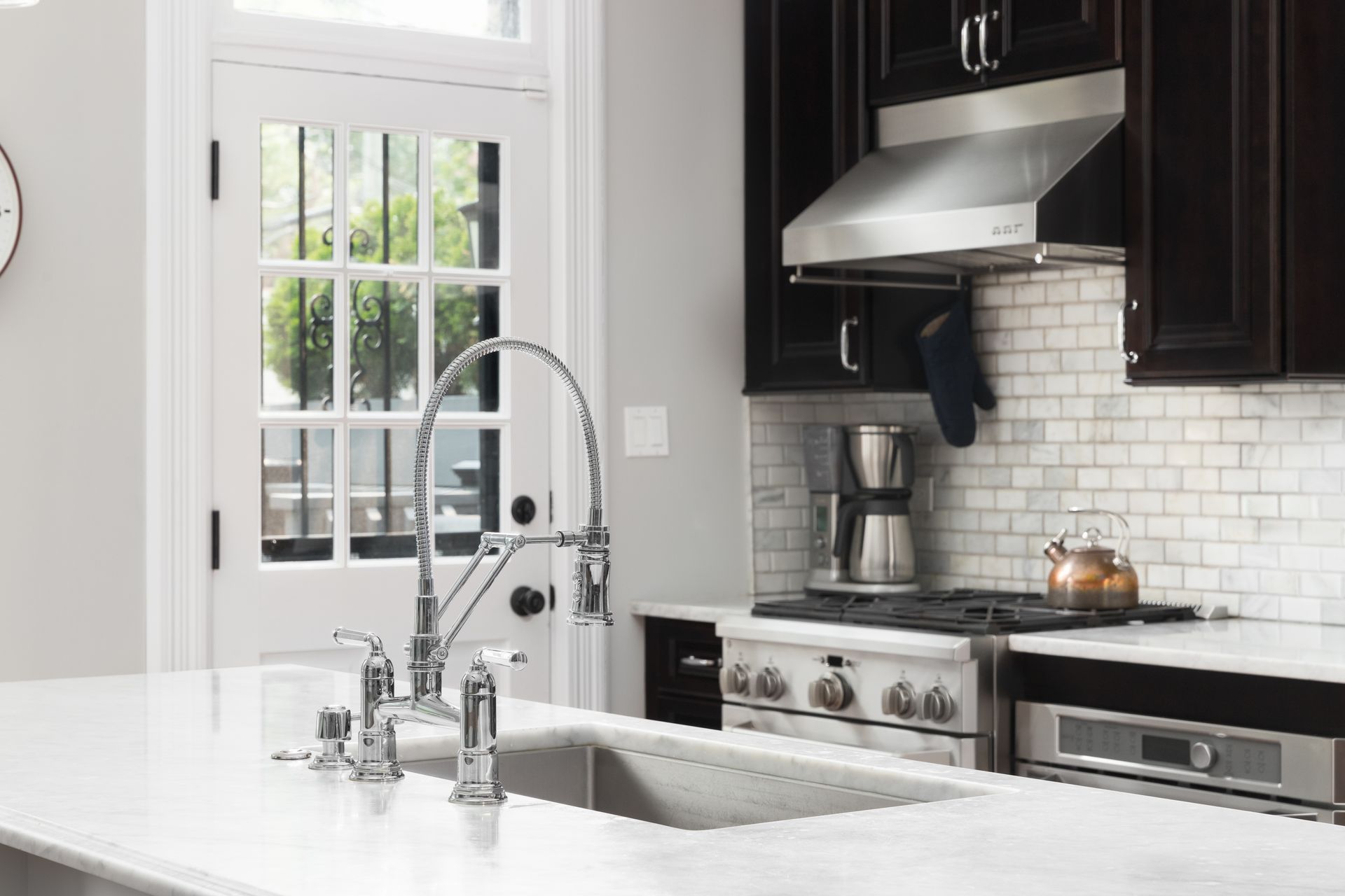A modern kitchen with dark cabinets, stainless steel appliances, and a marble countertop. A window and door provide natural light.