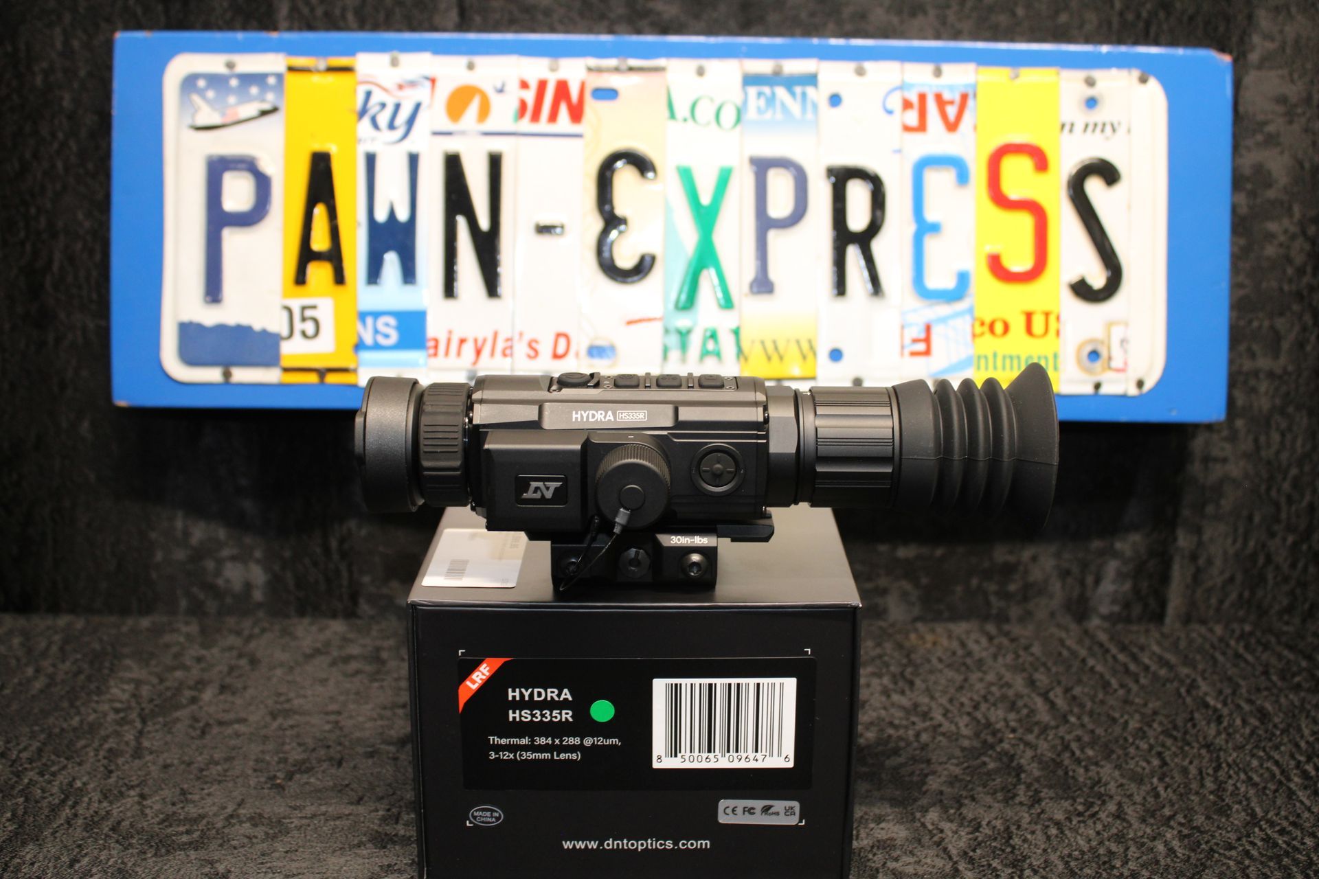 Blue license plate with “PAWN EXPRESS” mounted on a black device or box against a white background