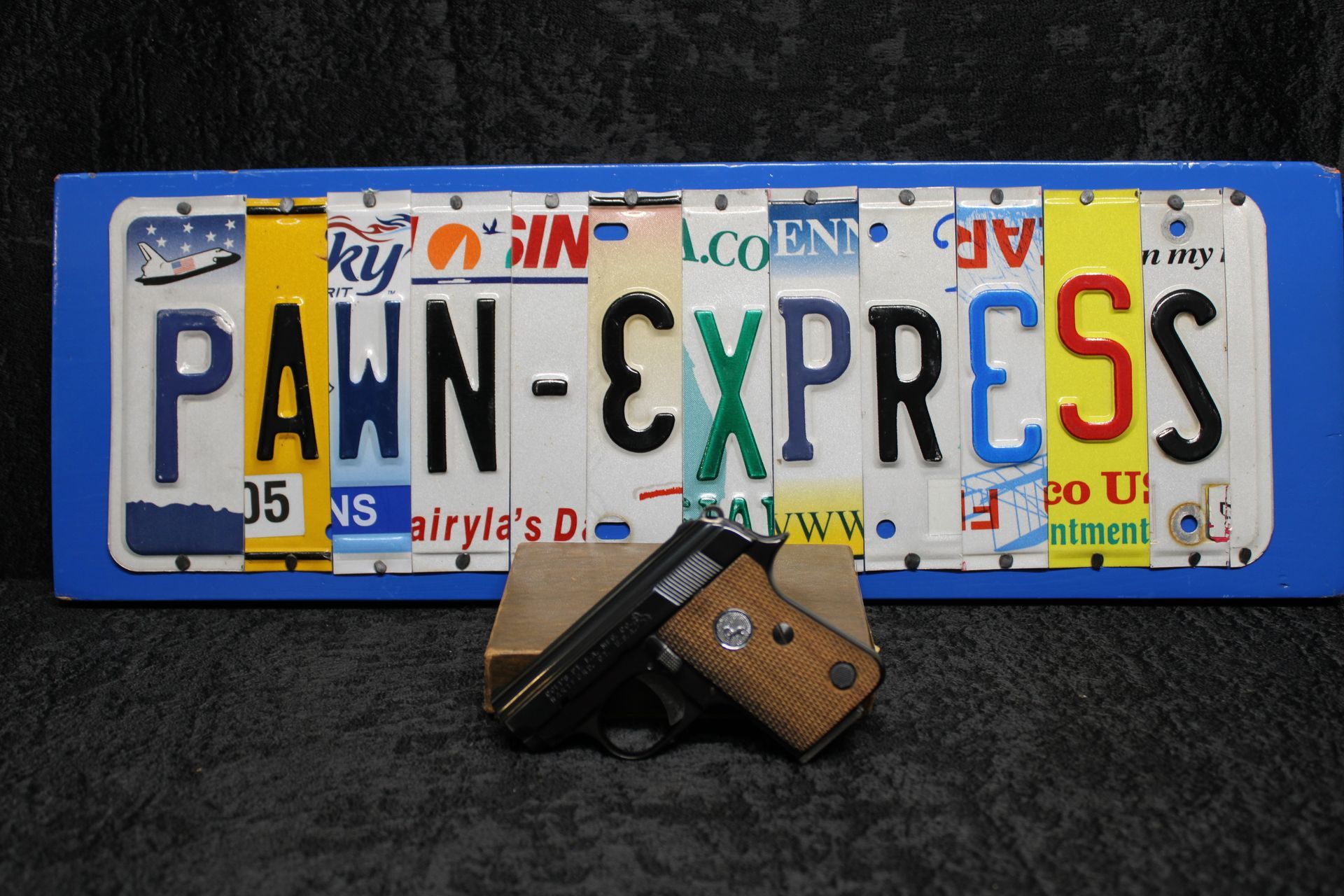 A compact handgun in front of a blue license plate reading “PAWN-EXPRESS.”