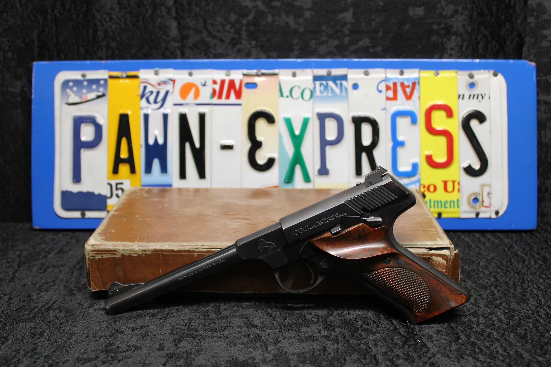 Handgun on a box in front of a Pawn Express sign and license plate background