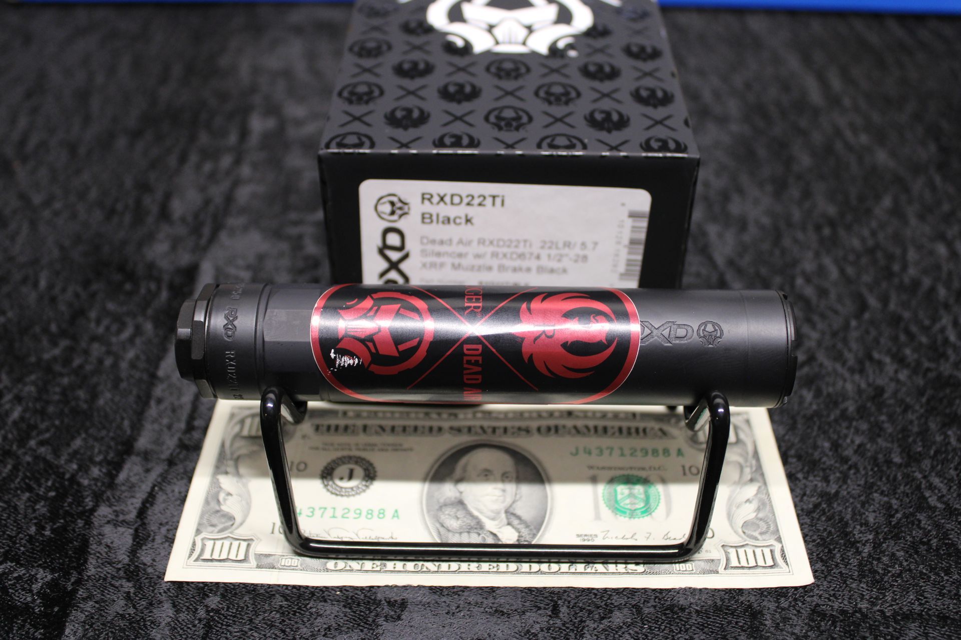 Black DX2TI battery-shaped device on a stand over a dollar bill in front of a black box.