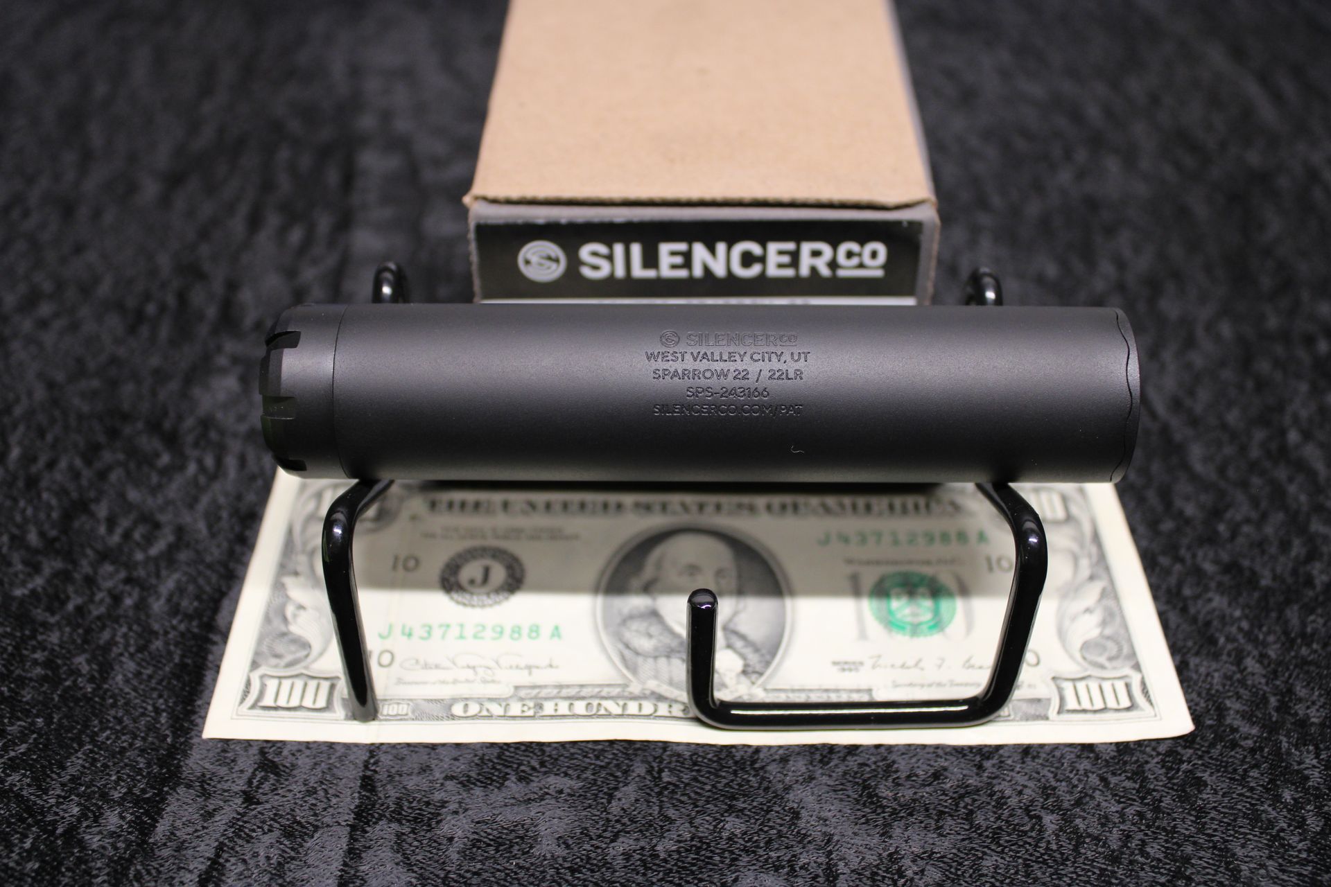Black silencer display on a $100 bill with SilencerCo logo and U-shaped stand