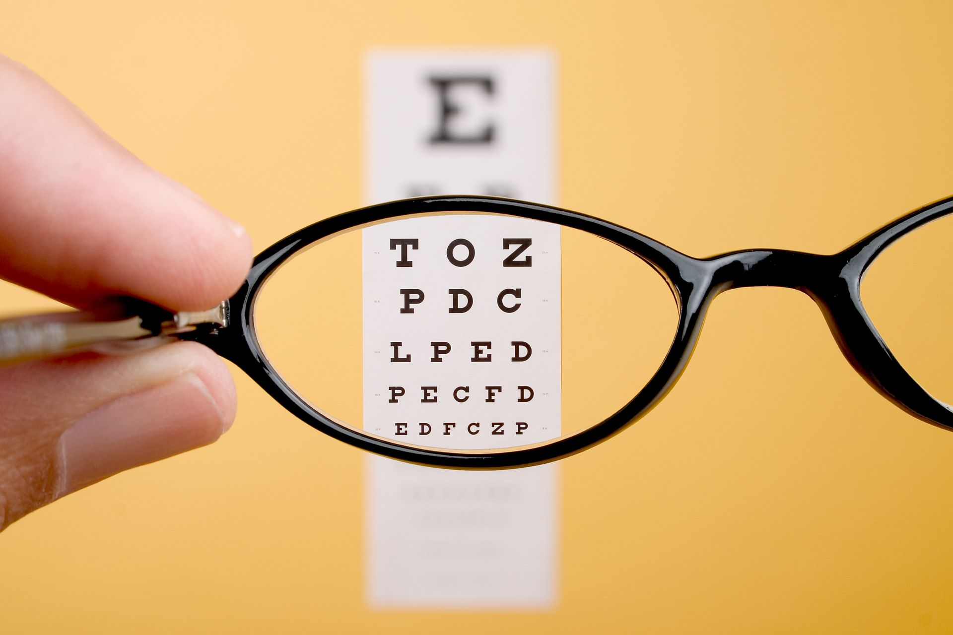 Hand holding eyeglasses in front of an eye chart, yellow background.