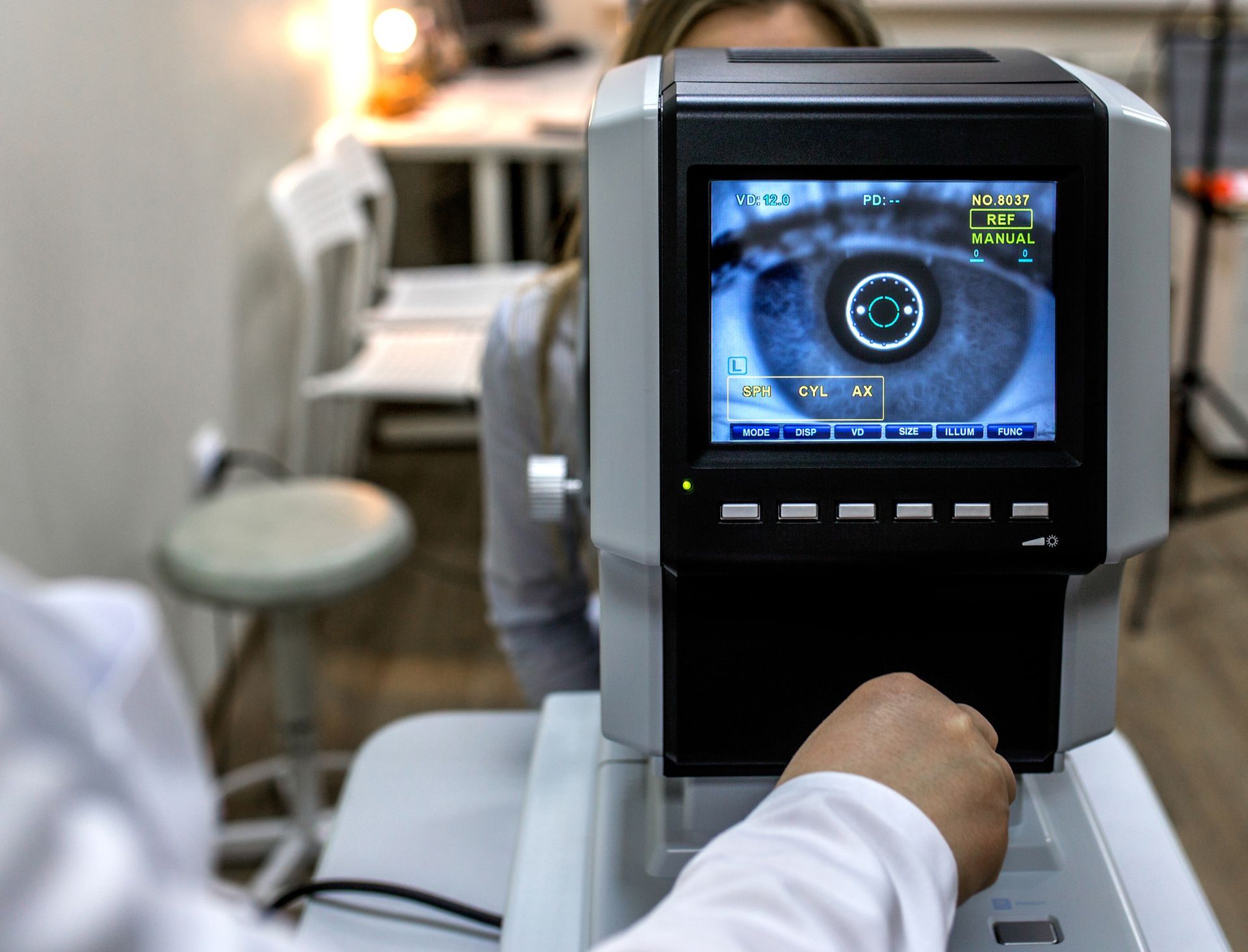 Eye exam in progress; patient's eye visible on a device screen. A hand operates the machine in a clinic setting.