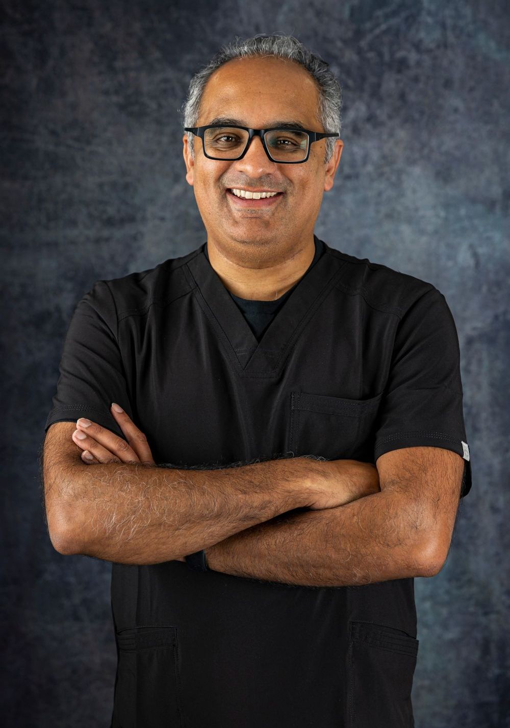 Dr Chavan in a black scrub top and glasses is standing with his arms crossed and smiling.