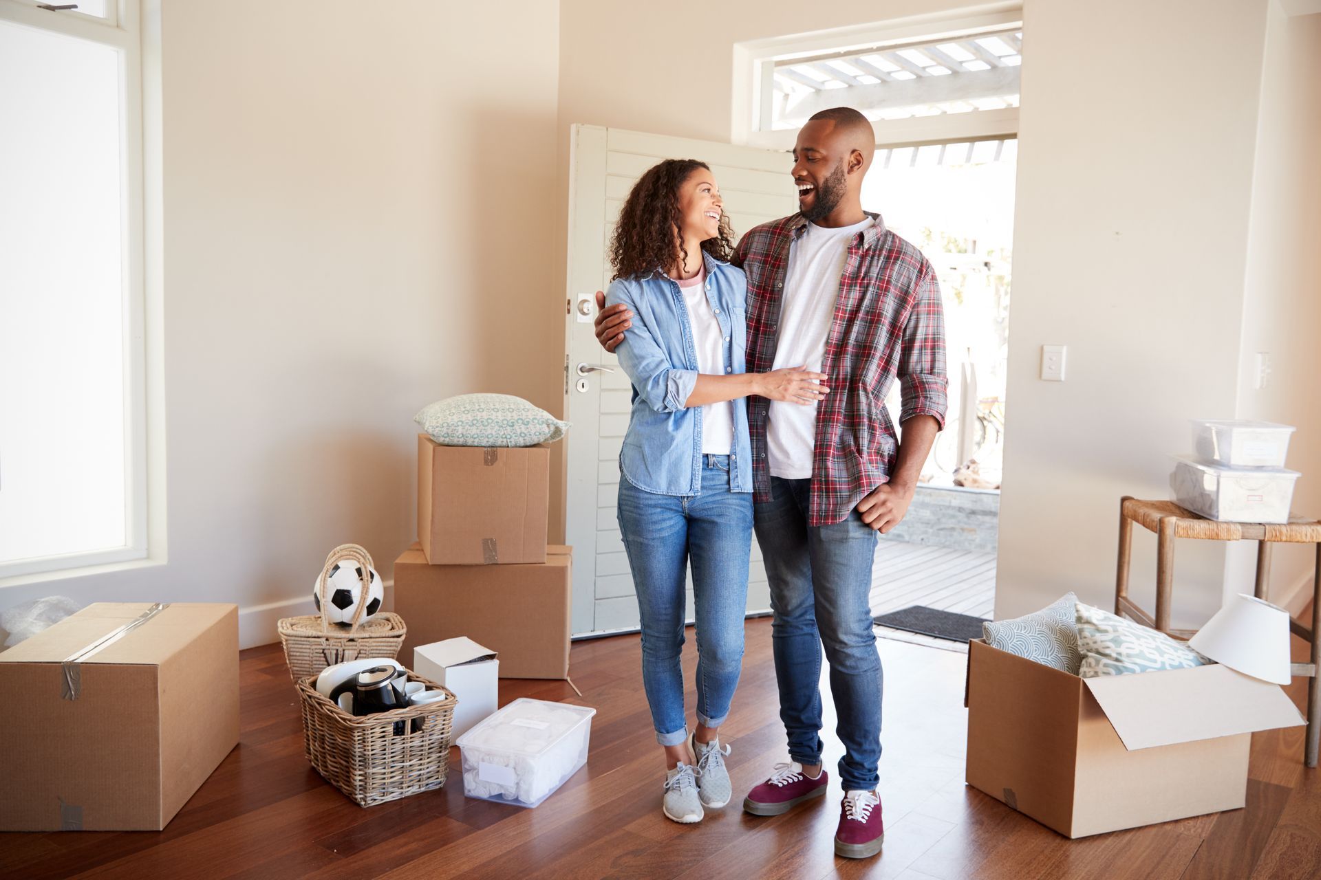Couple embraces in new home, surrounded by moving boxes, wooden floor, and open doorway.