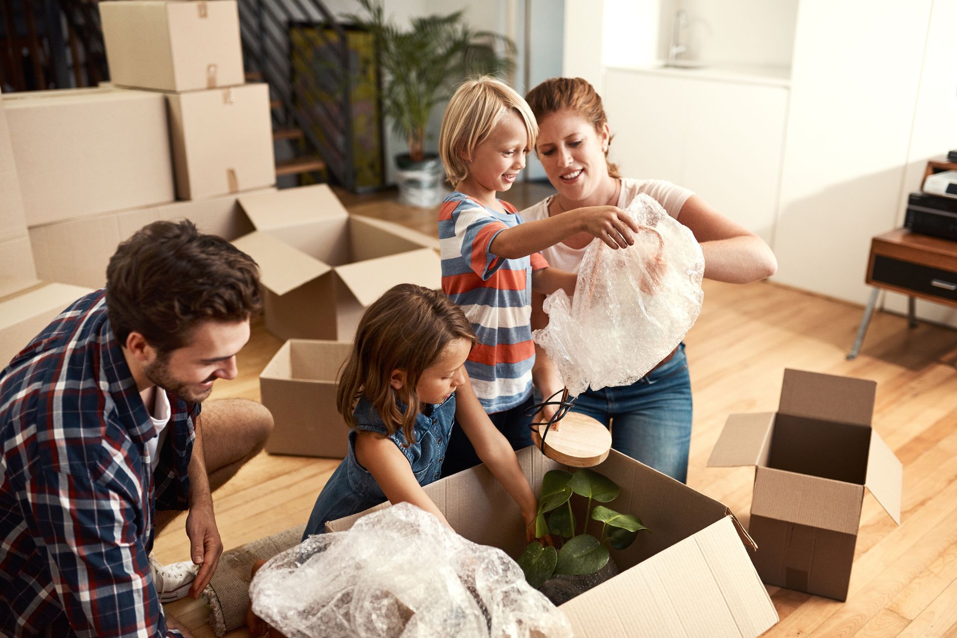 Family unpacking in new home, boxes and plants present. Woman and kids handling bubble wrap. Man smiles, wooden floors.
