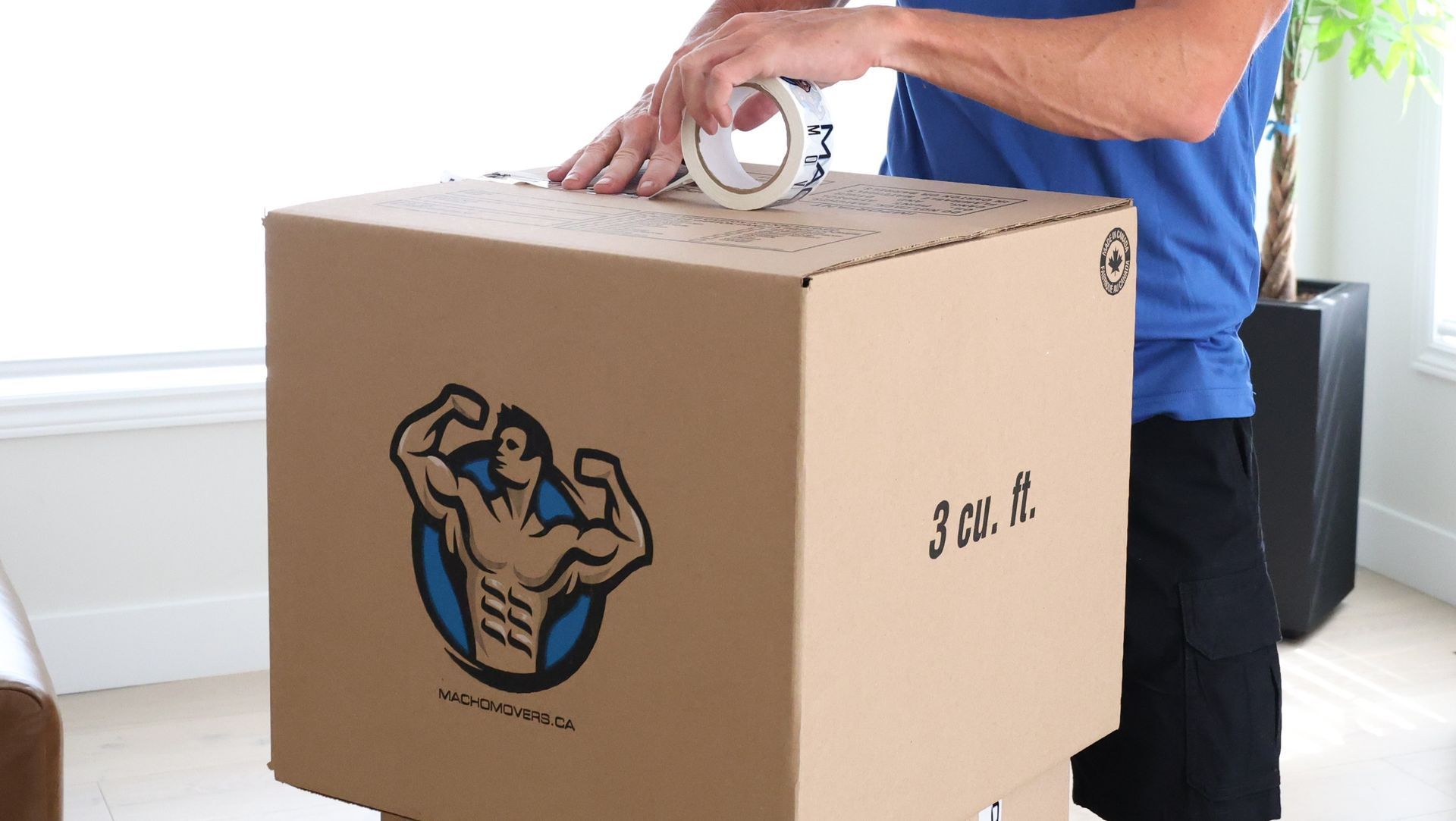 A person in a blue shirt is using a tape dispenser to seal a large cardboard shipping box marked with a muscular logo.