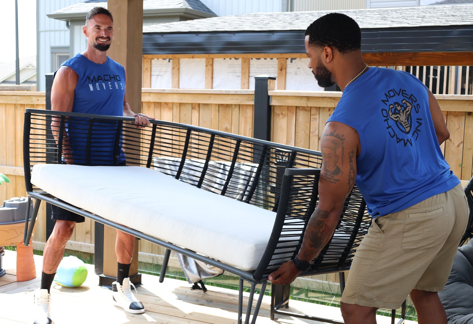 Two people wearing blue sleeveless shirts work together to move a black wicker outdoor sofa on a patio.