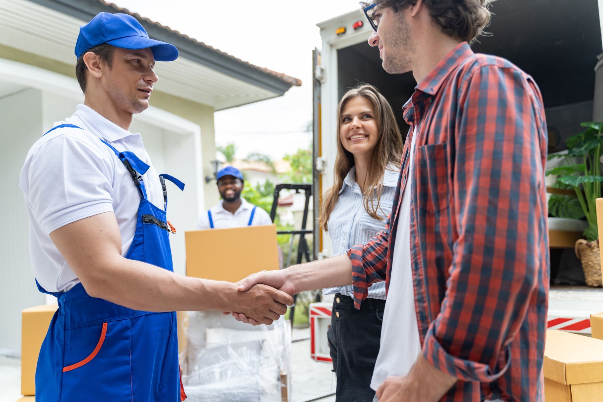 Movers shake hands with a couple at a home. Another mover and moving truck in the background.