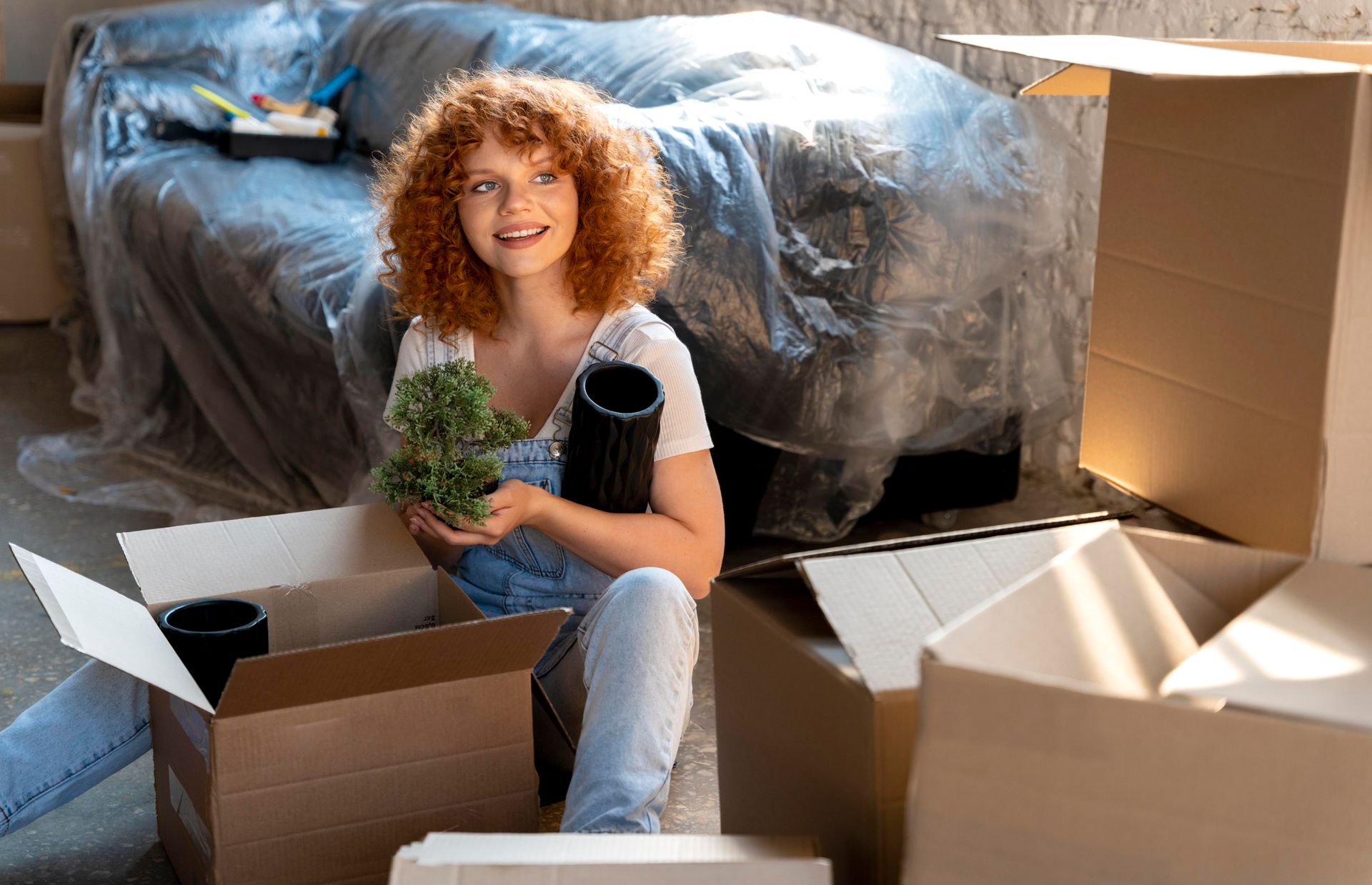 A woman is sitting on the floor in a living room surrounded by cardboard boxes holding a plant.