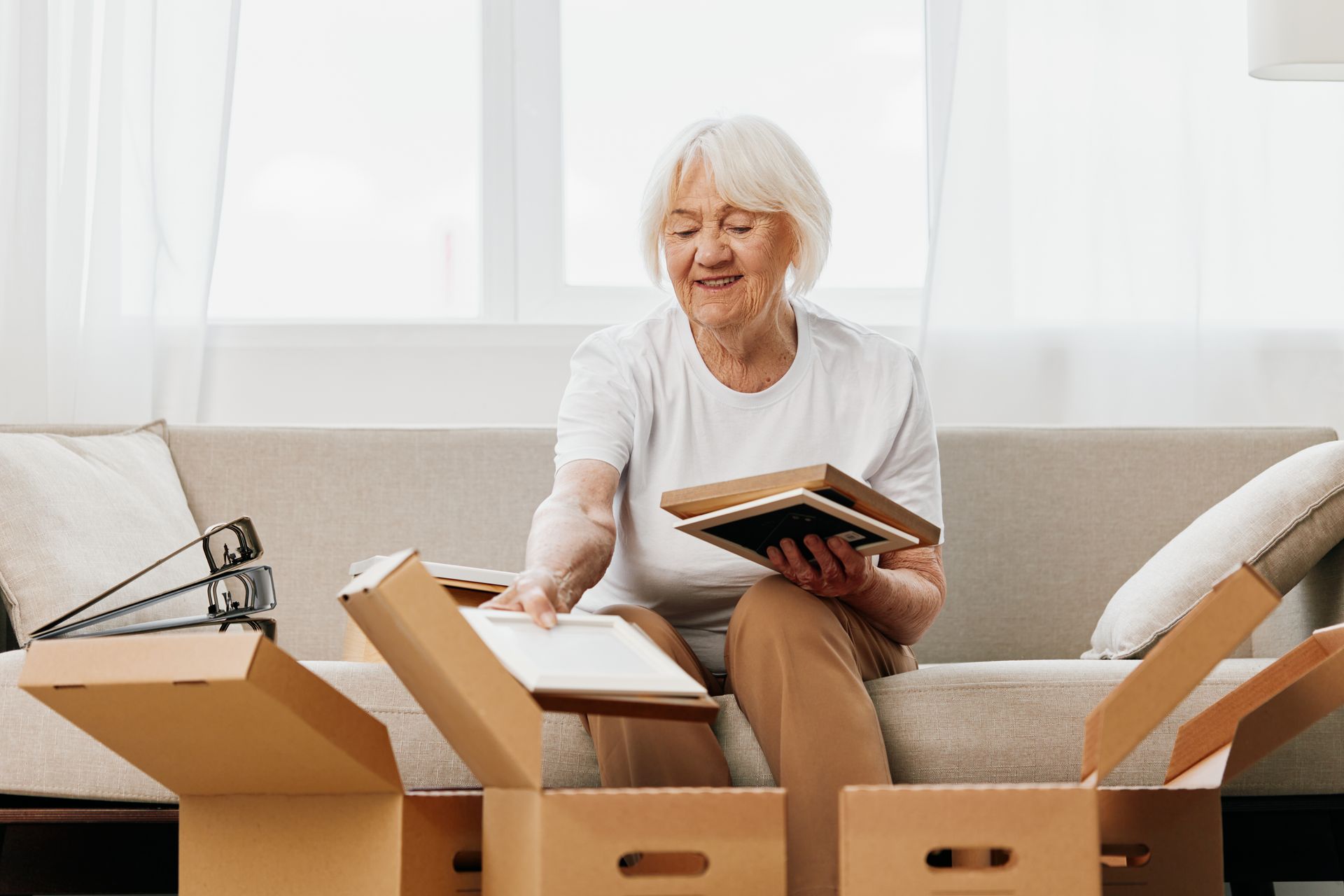 An elderly woman is sitting on a couch surrounded by cardboard boxes.