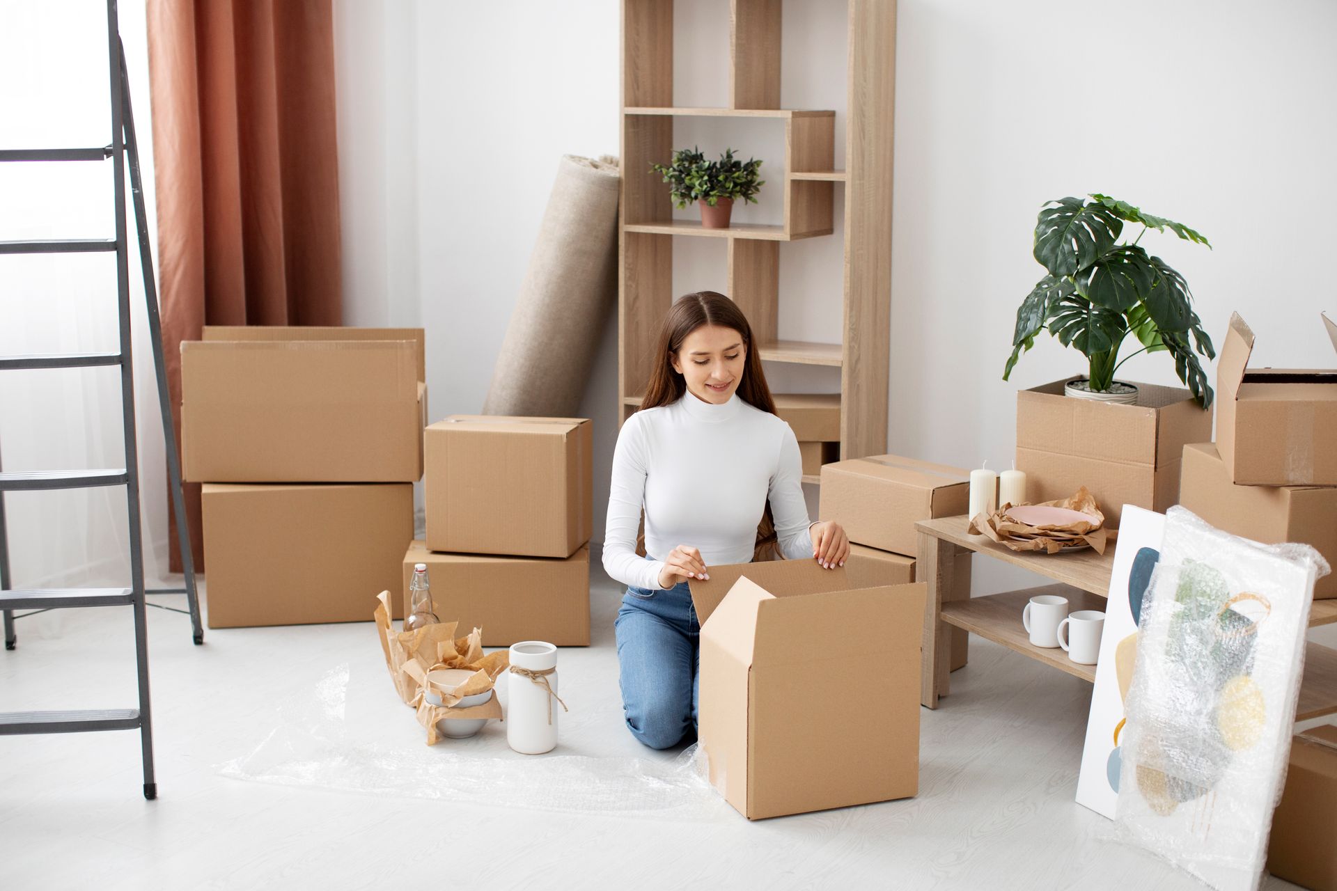A woman is kneeling on the floor in a room filled with cardboard boxes.