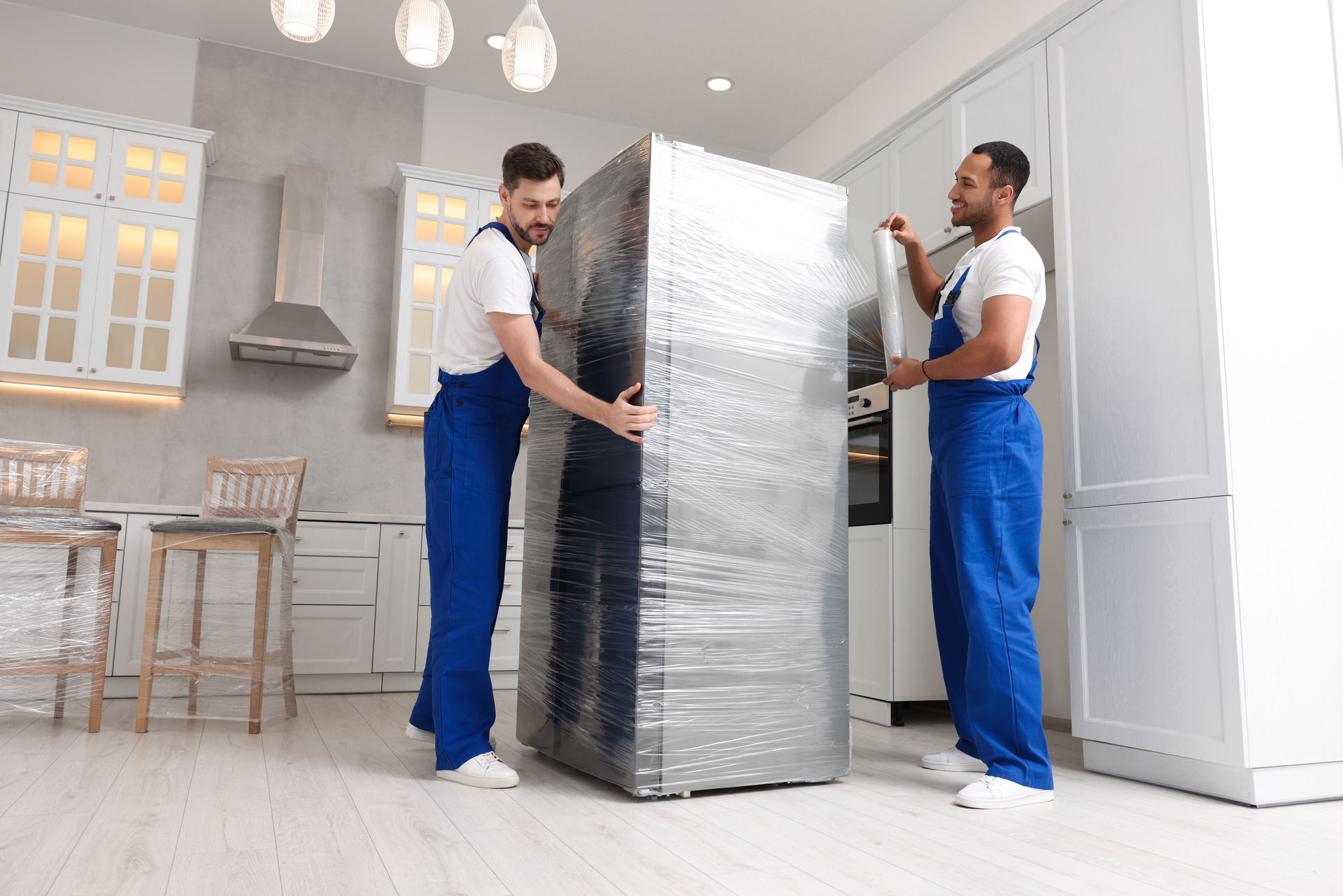Two men are wrapping a refrigerator in plastic wrap in a kitchen.