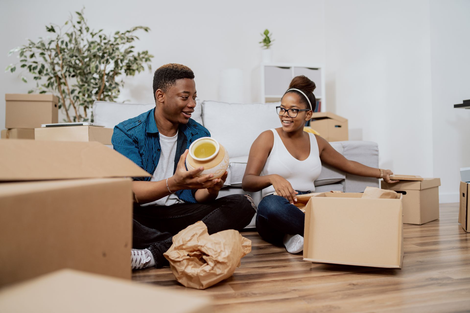 A man and a woman are sitting on the floor in a living room surrounded by cardboard boxes.