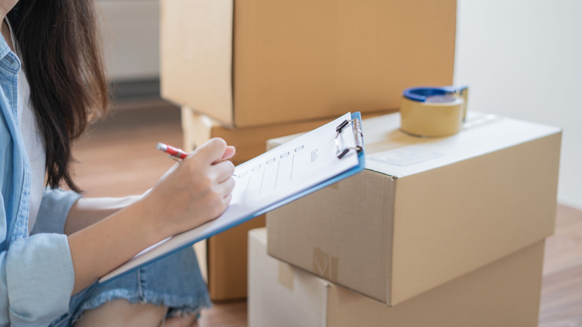 A woman is sitting in front of a pile of cardboard boxes and writing on a clipboard.