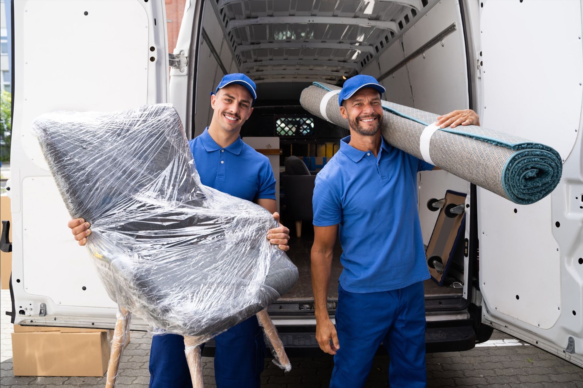Two movers are holding a chair and a rug in front of a van.