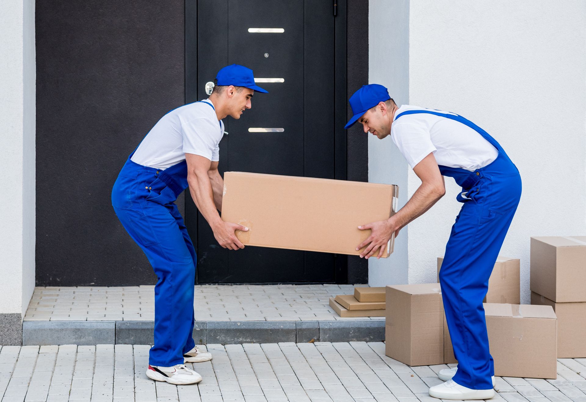 Two men in blue overalls are carrying a cardboard box.