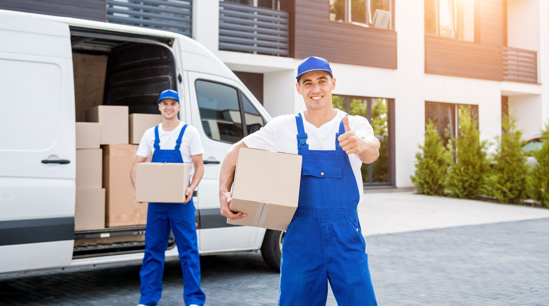 two movers are standing in front of a van holding boxes and giving a thumbs up .