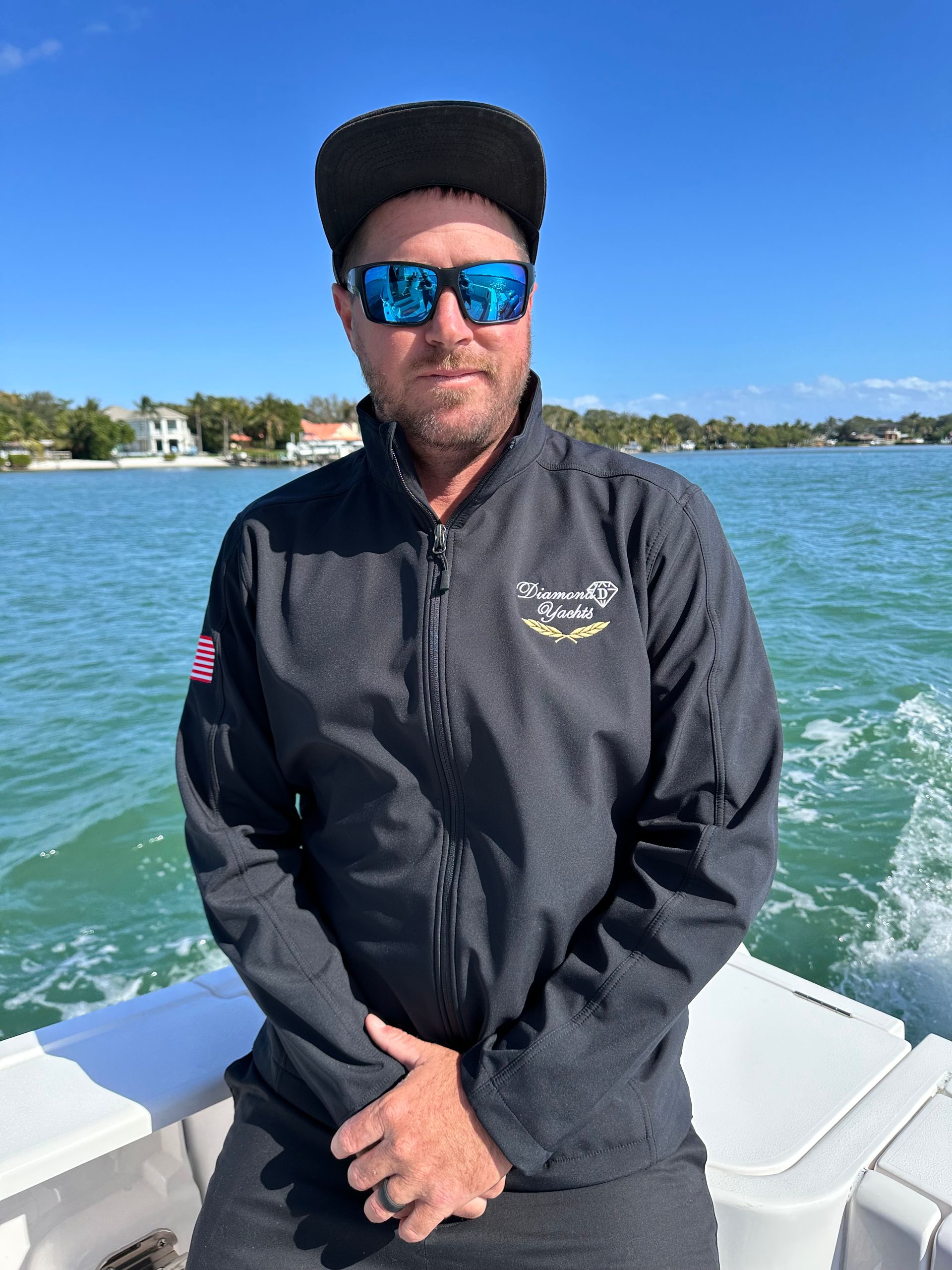 Man in sunglasses and black jacket on a boat, smiling, with water and houses in the background.