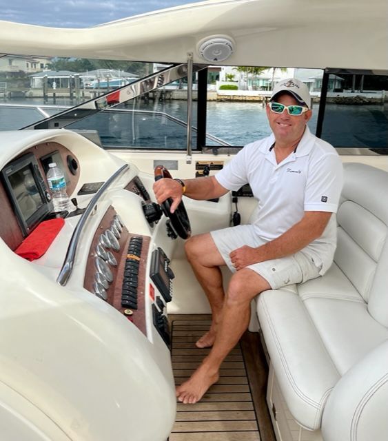 Man piloting a yacht, wearing a white shirt, shorts, and sunglasses. He is smiling in the vessel's cockpit.