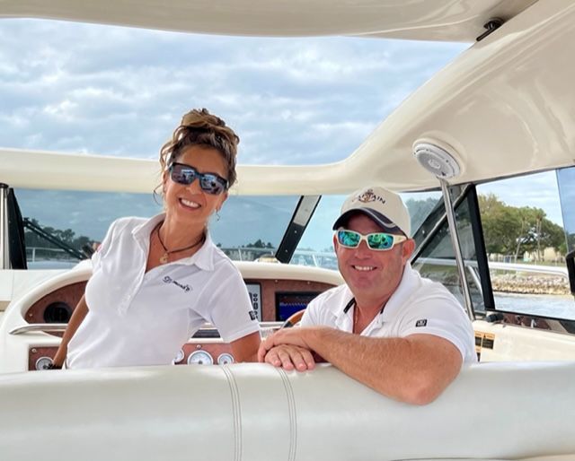 Two people smiling on a boat. Woman in white shirt and sunglasses, man in hat and sunglasses.
