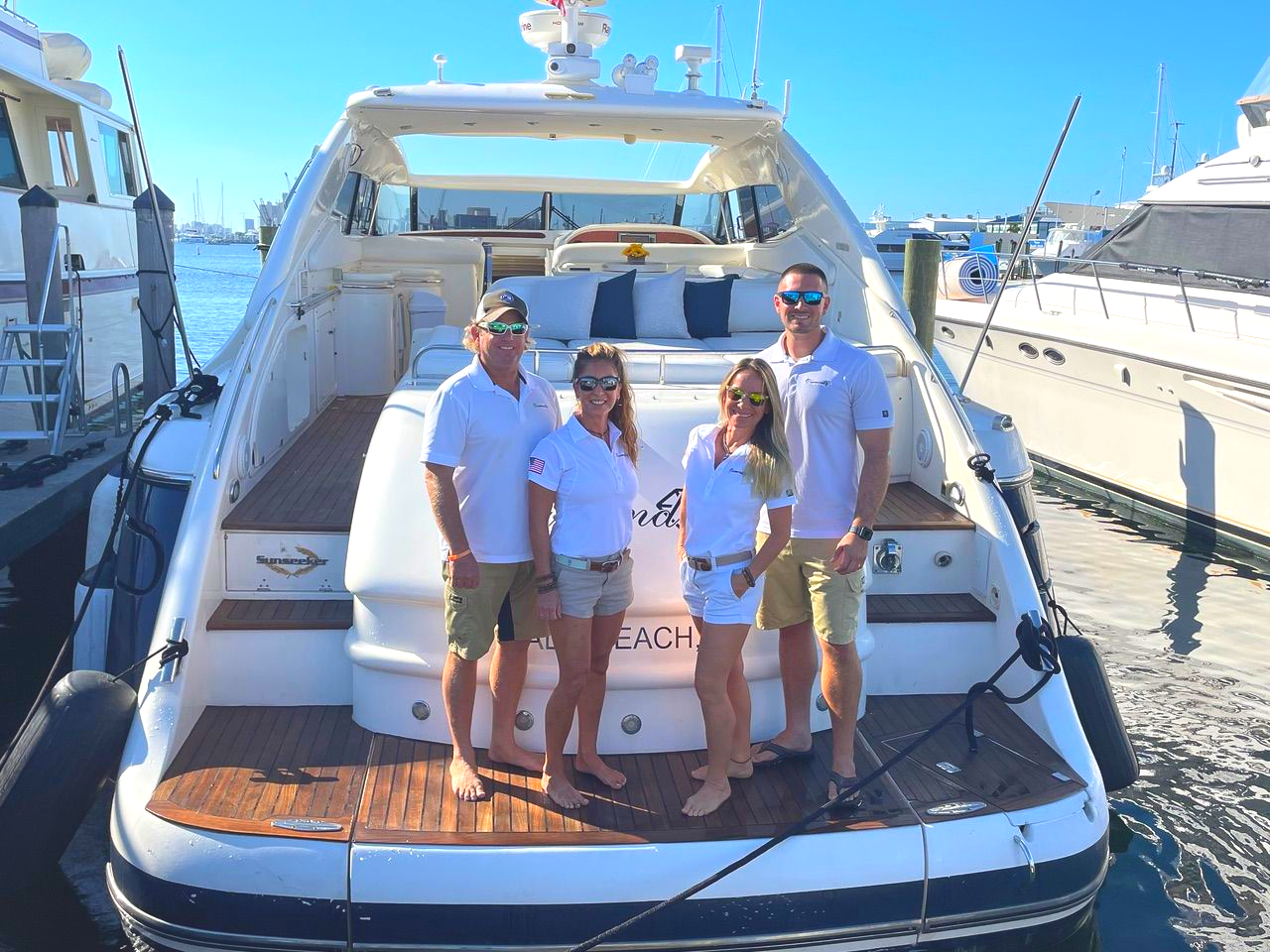 Four people pose on the back of a yacht in a harbor on a sunny day.