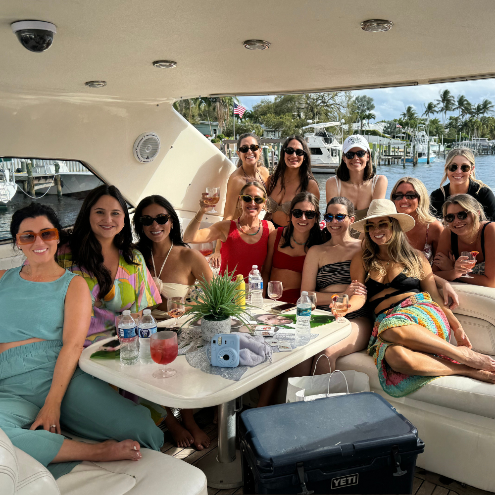 Group of women on a boat, sitting at a table with drinks and snacks. Sunshine, smiles, and relaxation.