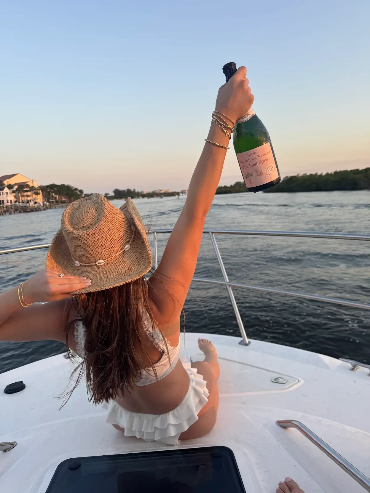 Woman on a boat raises a champagne bottle, celebrating. Wearing a straw hat and white bikini at sunset.